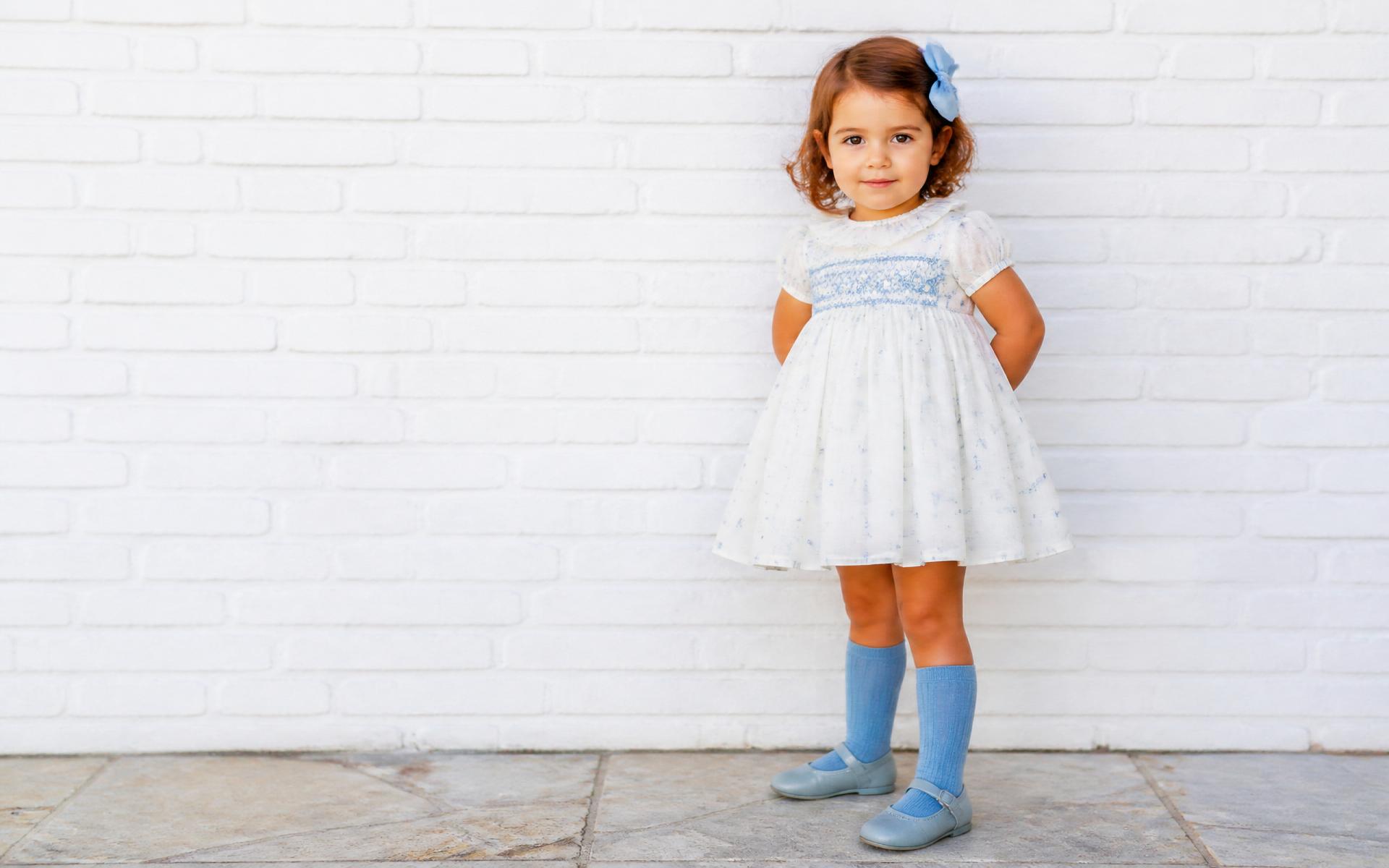 A toddler in a hand-smocked Spanish heirloom dress against a whitewashed brick wall